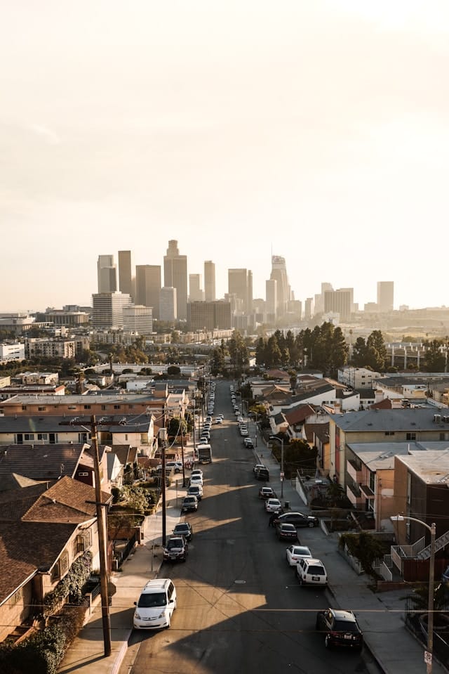 A city street lined with parked cars and houses leads toward a distant skyline of tall buildings under a hazy, sunlit sky.